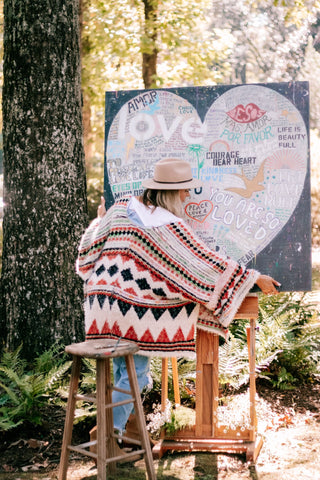 A woman painting a love heart on a canvas in the woods. Serene nature setting inspires artistic expression.